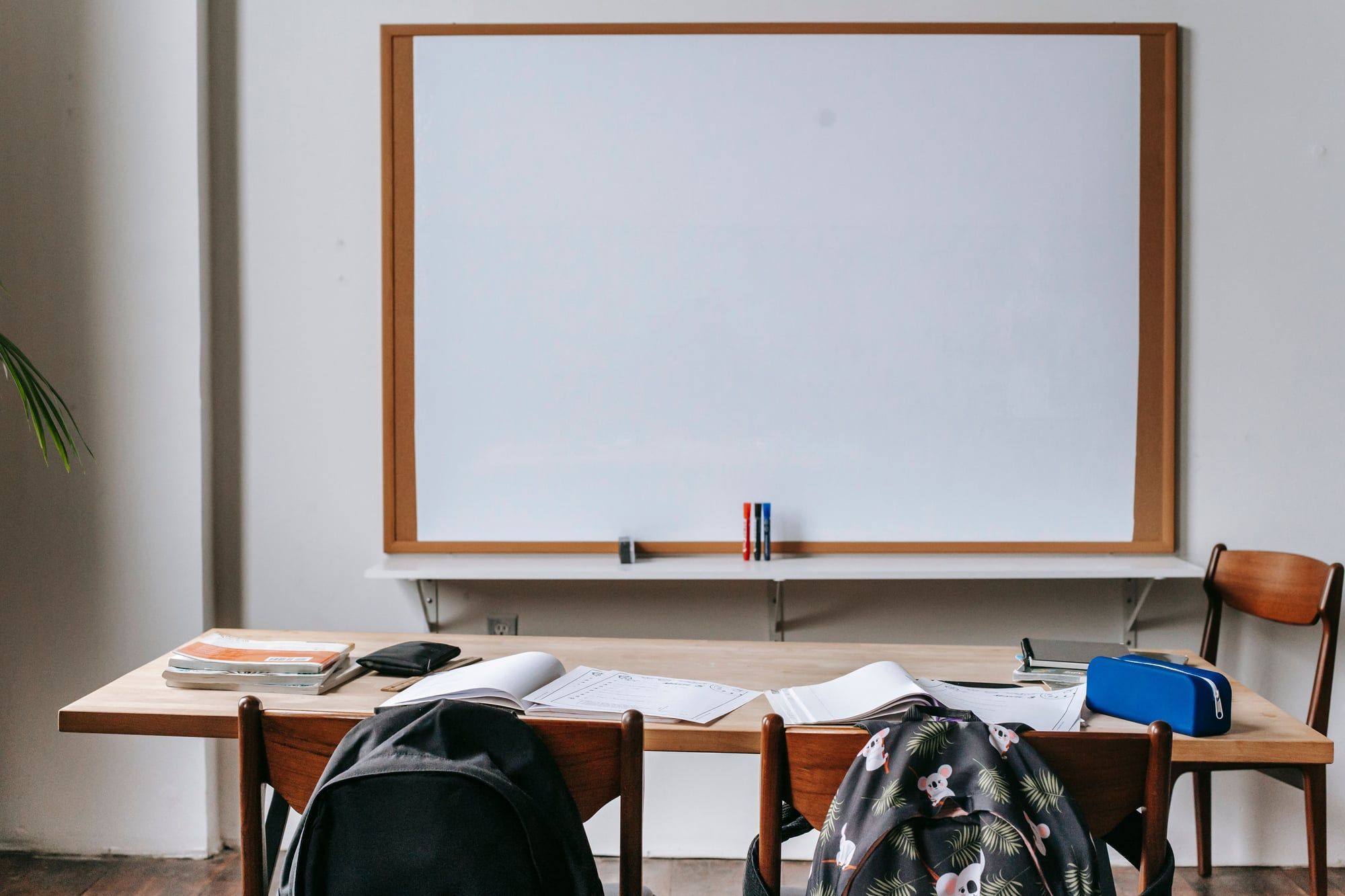 An empty classroom with a blank whiteboard, books on a wooden table and school bags hanging on the chairs